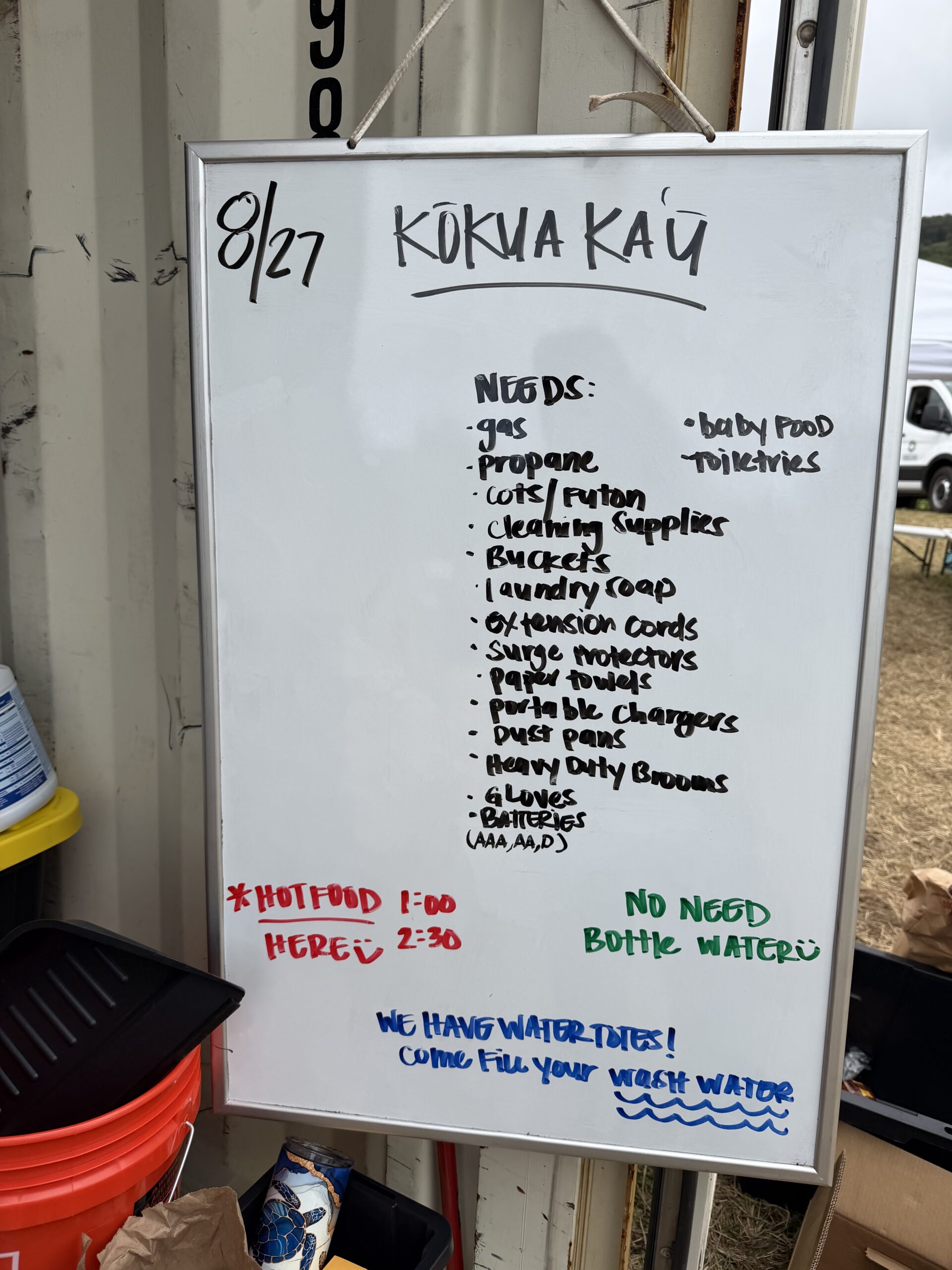 Handwritten whiteboard dated 8/21 headed Kokua Kau, listing community needs including canned food, hygiene products, propane, portable stoves, gas, baby items, paper goods, pet food and cleaning supplies.