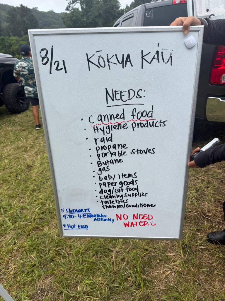 Handwritten whiteboard dated 8/21 headed Kokua Kau, listing community needs including canned food, hygiene products, propane, portable stoves, gas, baby items, paper goods, pet food and cleaning supplies.