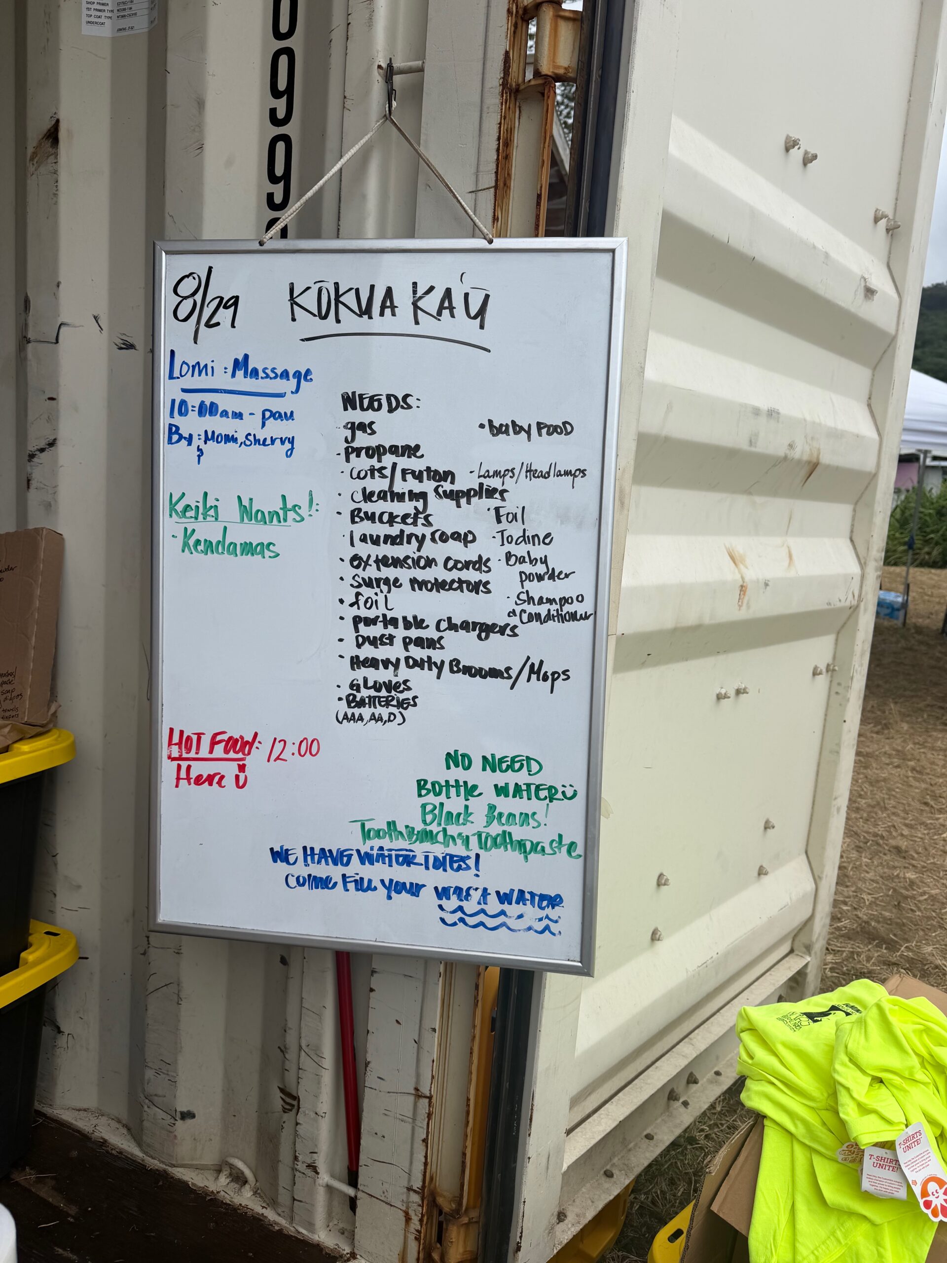 Handwritten whiteboard dated 8/21 headed Kokua Kau, listing community needs including canned food, hygiene products, propane, portable stoves, gas, baby items, paper goods, pet food and cleaning supplies.